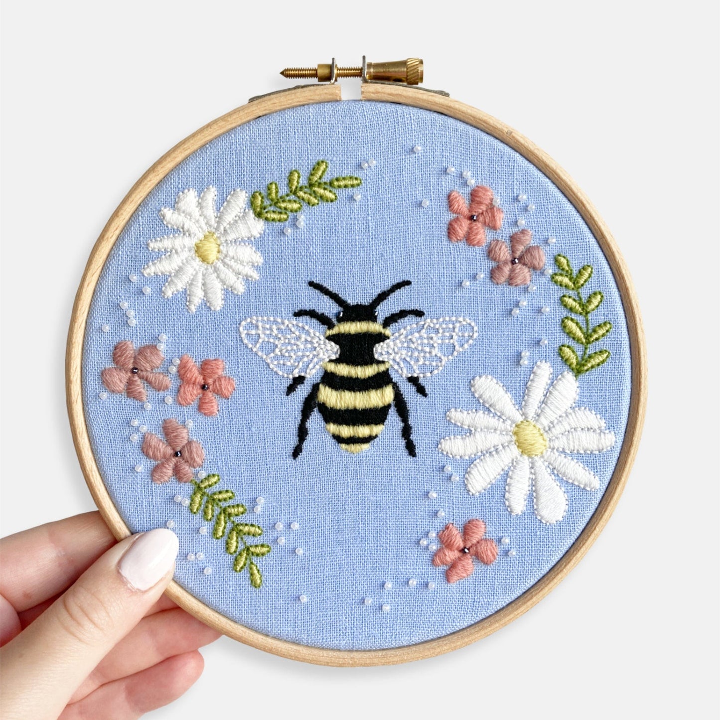 Floral Bee Embroidery Kit - Kirsty Freeman Design. A close up photograph of the embroidery kit in shades of pink, green, white and yellow, embroidered onto a sky blue fabric.