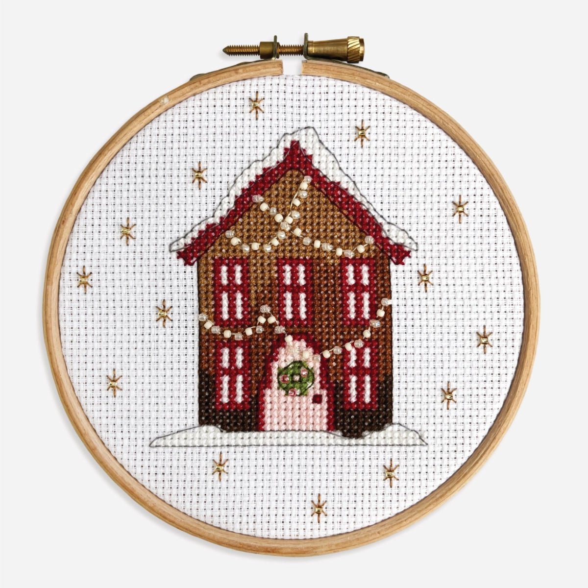Close-up of finished Gingerbread House cross stitch kit with red windows, a pink door and twinkly strings of fairy lights.