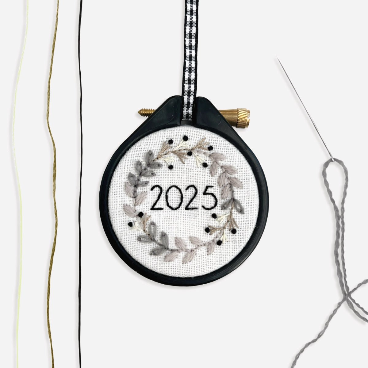 Completed Mini Christmas Wreath Embroidery Kit 2025 monochrome festive design next to cream, beige and black thread and a needle threaded with grey crewel wool.