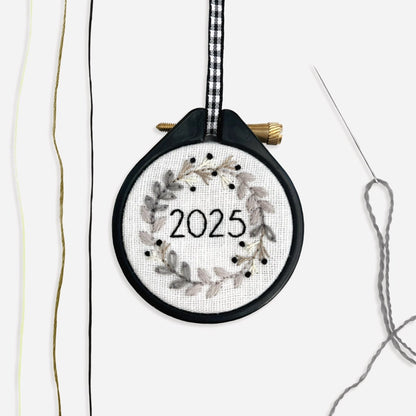 Completed Mini Christmas Wreath Embroidery Kit 2025 monochrome festive design next to cream, beige and black thread and a needle threaded with grey crewel wool.