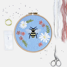 Load image into Gallery viewer, Bee Embroidery Kit - Kirsty Freeman Design. The bee is stitched in the centre of the blue linen fabric, and surrounded by an embroidered floral wreath of daisies and wildflowers. The design is presented inside an embroidery hoop frame, and is surrounded by materials included inside the kit.

