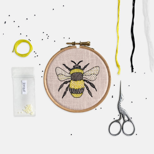 Bumble Bee Embroidery Kit - Kirsty Freeman Design. Modern embroidery hoop with bee emblem stitched onto pink linen fabric, surrounded by embroidery materials.