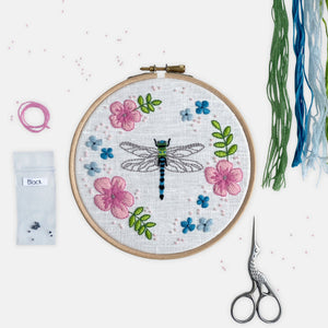 Dragonfly Embroidery Kit - Kirsty Freeman Design. A grey, blue and green dragonfly stitched onto white linen fabric, and surrounded by pink and blue flowers, green leaves and pink seed beads.