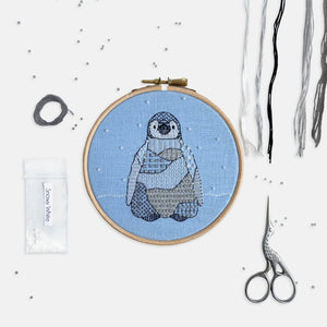 Penguin Embroidery Kit - Kirsty Freeman Design. A penguin stitched using modern, patterned embroidery stitches, created in black, white and shades of grey. The design is stitched onto blue linen fabric and has white seed bead snowflake stitched onto the background.