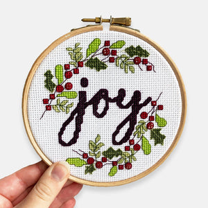 Joy Christmas Cross Stitch Kit - Kirsty Freeman Design. A festive embroidery hoop design, created using DMC stranded cotton and Toho seed beads. The design consists of berries, holly, mistletoe and leaves, surrounding the word 'joy', using Christmas colours in shades of red and green.